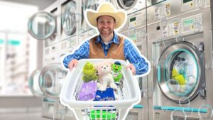 Smiling man in a cowboy hat and vest pushes a laundry basket filled with stuffed animals and laundry supplies in a laundromat.