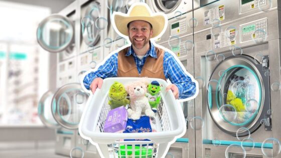 Smiling man in a cowboy hat and vest pushes a laundry basket filled with stuffed animals and laundry supplies in a laundromat.