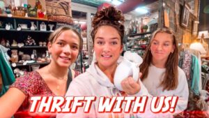 Three friends in a cluttered thrift store; center holds a white vase, bottom text 'THRIFT WITH US!'