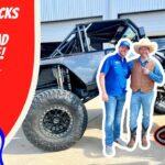 Two men in cowboy hats pose with a huge lifted off-road truck outside a garage, front and center, with bold red branding on the left and logos nearby.