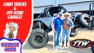 Two men in cowboy hats pose with a huge lifted off-road truck outside a garage, front and center, with bold red branding on the left and logos nearby.