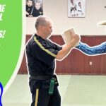 Playful thumbnail showing a martial arts instructor in black gi and a cowboy in a hat sparring with a wooden board in a kids' martial arts video thumbnail.