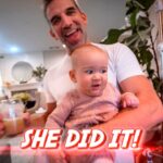 Father joyfully holding a smiling baby indoors, with a bold caption reading 'SHE DID IT!' in the foreground.