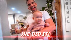 Father joyfully holding a smiling baby indoors, with a bold caption reading 'SHE DID IT!' in the foreground.
