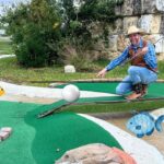 Man in a cowboy hat kneels on a mini-golf green, pointing at a white ball with a putter; cartoon fish decorate the course.