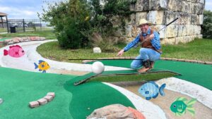 Man in a cowboy hat kneels on a mini-golf green, pointing at a white ball with a putter; cartoon fish decorate the course.