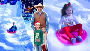 Smiling man in a cowboy hat and vest poses with two children at a snowy indoor slide, while others sled nearby under snowflakes.