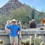 Promotional cutout of a man in a blue shirt with two children posing against a desert park backdrop with cacti, while a real cougar walks along a low wall nearby at Pinnacle Peak Park.