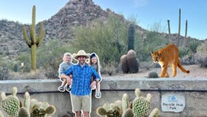 Promotional cutout of a man in a blue shirt with two children posing against a desert park backdrop with cacti, while a real cougar walks along a low wall nearby at Pinnacle Peak Park.