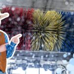 Man in a cowboy hat and brown leather vest points at a large colorful car-wash brush, with soap bubbles in the foreground.