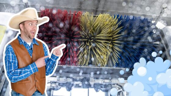 Man in a cowboy hat and brown leather vest points at a large colorful car-wash brush, with soap bubbles in the foreground.