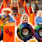 Man in a cowboy hat and blue plaid shirt stands beside rainbow-colored inflatable tires with two children in a bright trampoline park.