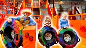 Man in a cowboy hat and blue plaid shirt stands beside rainbow-colored inflatable tires with two children in a bright trampoline park.