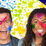 Two smiling people with colorful paint splatters on their faces against a bright splattered backdrop