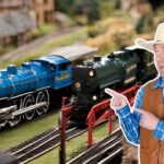 Man in a cowboy hat and brown vest points at blue and green model locomotives on a miniature railroad track set.