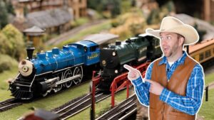 Man in a cowboy hat and brown vest points at blue and green model locomotives on a miniature railroad track set.
