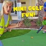 Two people on a mini golf green: a surprised woman in a yellow safety vest watches as a worker in a blue helmet uses a tool toward a small orange ball, with 'MINI GOLF FUN!' text overlay.