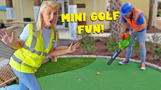 Two people on a mini golf green: a surprised woman in a yellow safety vest watches as a worker in a blue helmet uses a tool toward a small orange ball, with 'MINI GOLF FUN!' text overlay.