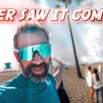 Bearded man on a sunny beach wearing teal shirt and reflective sunglasses, taking a selfie with smoke and two people in the background and bold red text overlay saying 'NEVER SAW IT COMING!'
