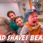Crying baby in orange overalls held by an adult, while three older siblings gaze on; bold red text at the bottom reads 'DAD SHAVES BEARD'.