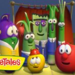 Group of VeggieTales vegetable characters posing together on a stage with a microphone and red curtain, smiling at the camera.