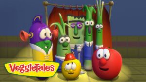 Group of VeggieTales vegetable characters posing together on a stage with a microphone and red curtain, smiling at the camera.