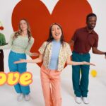 Group of five friends dancing in a bright studio with a large red heart backdrop and retro 'Love' lettering in the foreground; colorful 70s outfits.