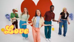 Group of five friends dancing in a bright studio with a large red heart backdrop and retro 'Love' lettering in the foreground; colorful 70s outfits.