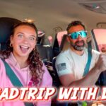 Woman in a pink hoodie smiles excitedly in the passenger seat as a bearded man drives wearing blue sunglasses; bold 'ROADTRIP WITH DAD' text overlays the image.