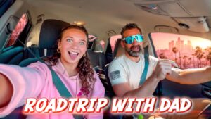 Woman in a pink hoodie smiles excitedly in the passenger seat as a bearded man drives wearing blue sunglasses; bold 'ROADTRIP WITH DAD' text overlays the image.