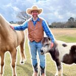 Man in a cowboy hat and vest standing with a large light-brown horse and a small pony in a grassy field, with a duck nearby.
