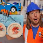Construction worker in an orange vest with blue helmet giving a thumbs up, holding a blue drawing and homemade 'goggles' made from paper plates, in a workshop setting.