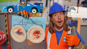 Construction worker in an orange vest with blue helmet giving a thumbs up, holding a blue drawing and homemade 'goggles' made from paper plates, in a workshop setting.