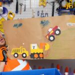 Man in a blue hard hat and orange safety vest holds up a cardboard craft featuring a yellow dump truck, a yellow excavator, and a red monster truck climbing a grassy hill in a workshop setting.