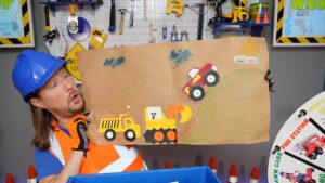 Man in a blue hard hat and orange safety vest holds up a cardboard craft featuring a yellow dump truck, a yellow excavator, and a red monster truck climbing a grassy hill in a workshop setting.