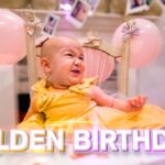 Baby in a yellow dress sits in a small chair at a birthday party with pink balloons in the background and 'GOLDEN BIRTHDAY!' text at the bottom