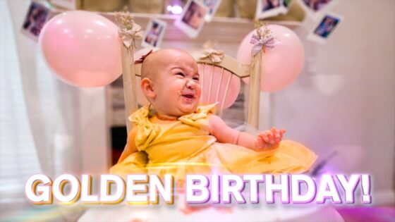 Baby in a yellow dress sits in a small chair at a birthday party with pink balloons in the background and 'GOLDEN BIRTHDAY!' text at the bottom