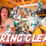 Woman gesturing in a cluttered basement full of boxes and junk, with a bold 'SPRING CLEAN' banner across the bottom.