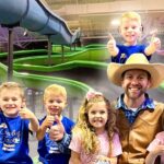 Man in a cowboy hat and brown vest posing with six kids in a colorful indoor water park, all giving thumbs up.