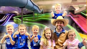Man in a cowboy hat and brown vest posing with six kids in a colorful indoor water park, all giving thumbs up.