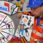 Construction worker in an orange safety vest and blue helmet holds a tool-filled coloring sheet beside a large wheel game titled with construction images.