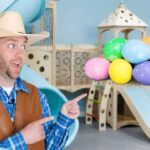 Man in a cowboy hat and brown vest points at a cluster of colorful Easter eggs indoors playground backdrop.