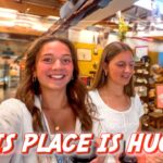 Two young women take a selfie in a busy, colorful gift shop with shelves full of items behind them.