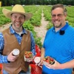 Two men smile in a strawberry field, one in a straw hat and leather vest, the other in a blue striped polo, holding strawberry containers and a jar of jam.