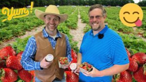 Two men smile in a strawberry field, one in a straw hat and leather vest, the other in a blue striped polo, holding strawberry containers and a jar of jam.