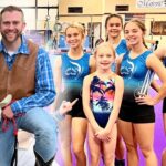 Man in a brown vest and blue plaid shirt sits with a white dog wearing a cowboy hat, posing with six young gymnasts in blue leotards in a gym with purple mats.