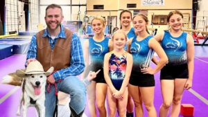 Man in a brown vest and blue plaid shirt sits with a white dog wearing a cowboy hat, posing with six young gymnasts in blue leotards in a gym with purple mats.