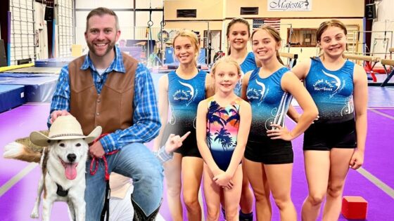 Man in a brown vest and blue plaid shirt sits with a white dog wearing a cowboy hat, posing with six young gymnasts in blue leotards in a gym with purple mats.