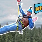 Man ziplining over a forest, wearing a blue helmet, brown vest and safety harness, with a 'WHOA!' cartoon bubble beside him.