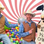 Two men in cowboy vests and hats pose in a colorful ball pit with a life-size cow mascot holding a sign that says 'EAT MOR CHIKIN' against a pink swirl background.
